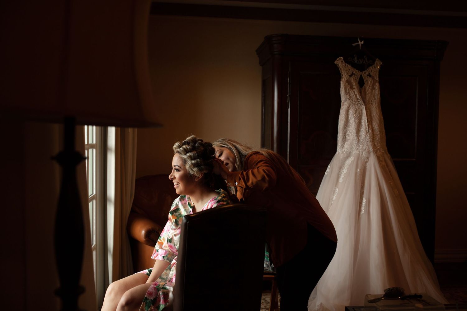 Bride sitting by window while bridesmaid adjusts her hair with wedding gown hanging nearby.