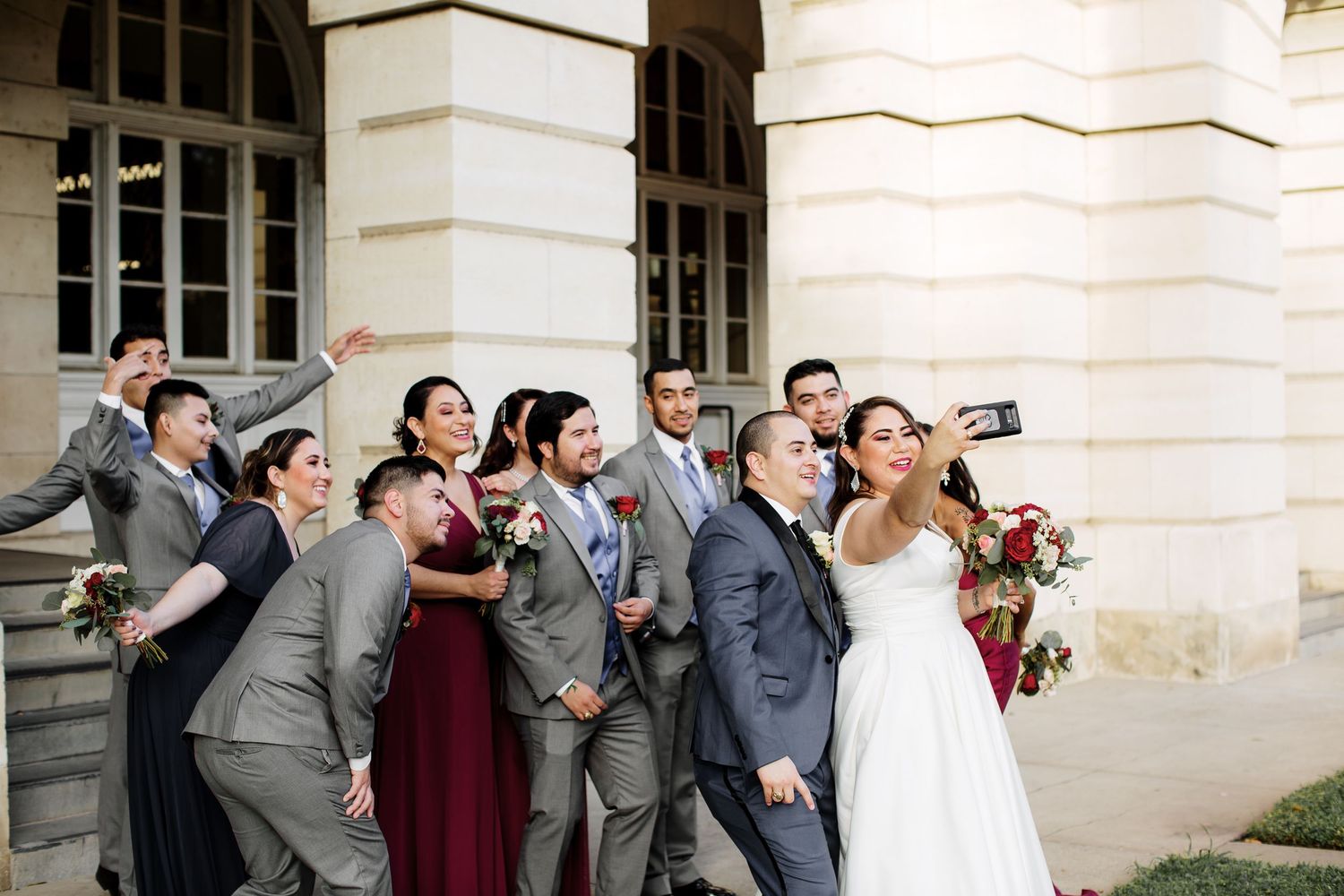 Wedding party posing playfully on steps of a grand stone building with bride and groom.