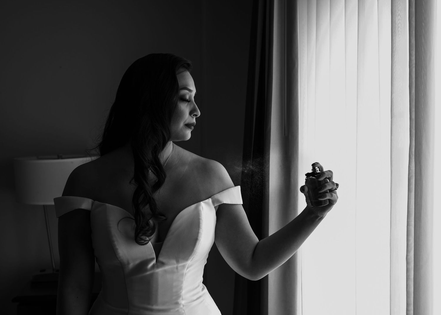 Elegant bride in off-shoulder gown spraying perfume by window in black and white.