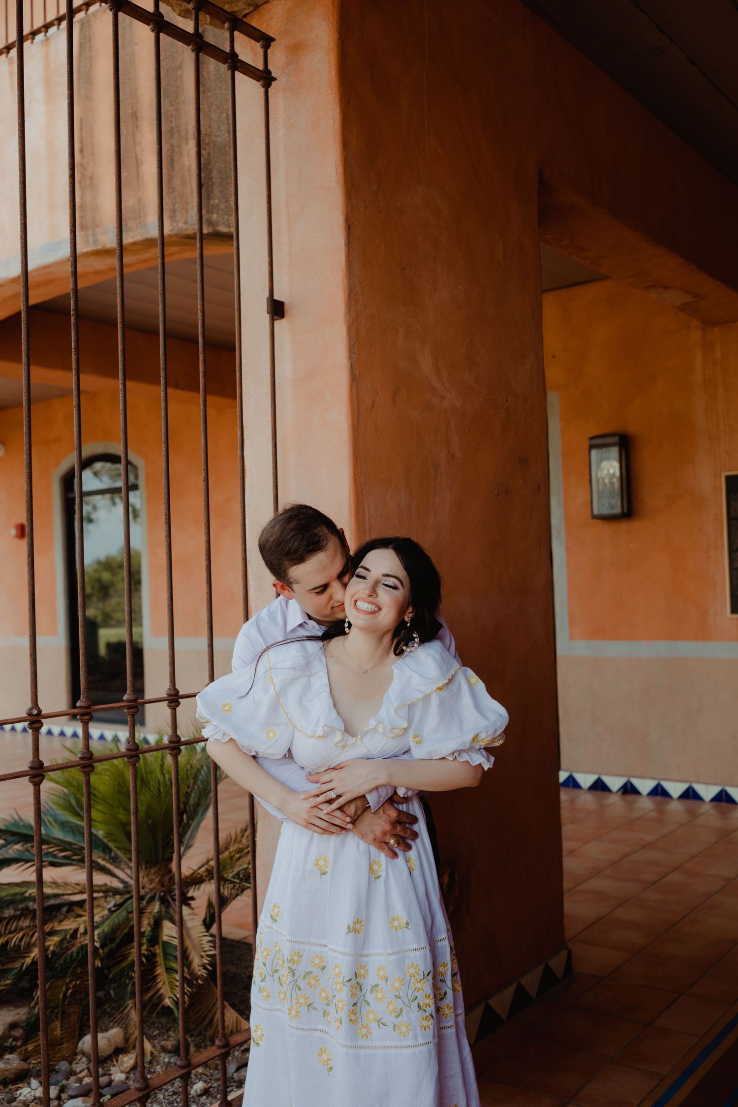 Couple embracing and smiling in front of a rustic adobe building with iron gate and colorful tilework.
