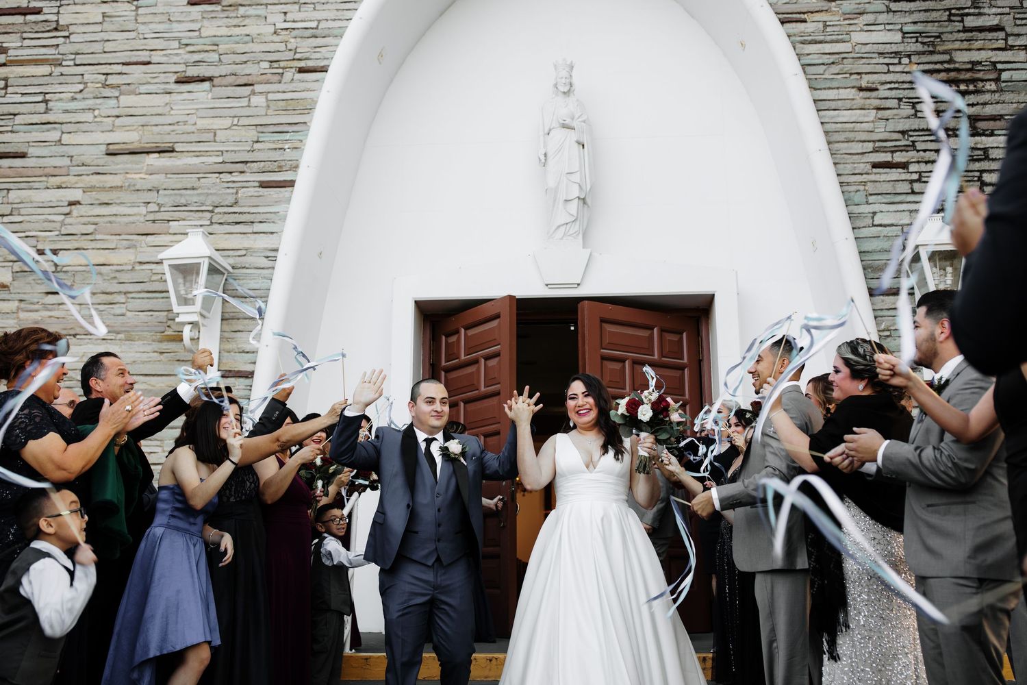 Bride and groom exit white stone church surrounded by cheering guests waving ribbon wands.