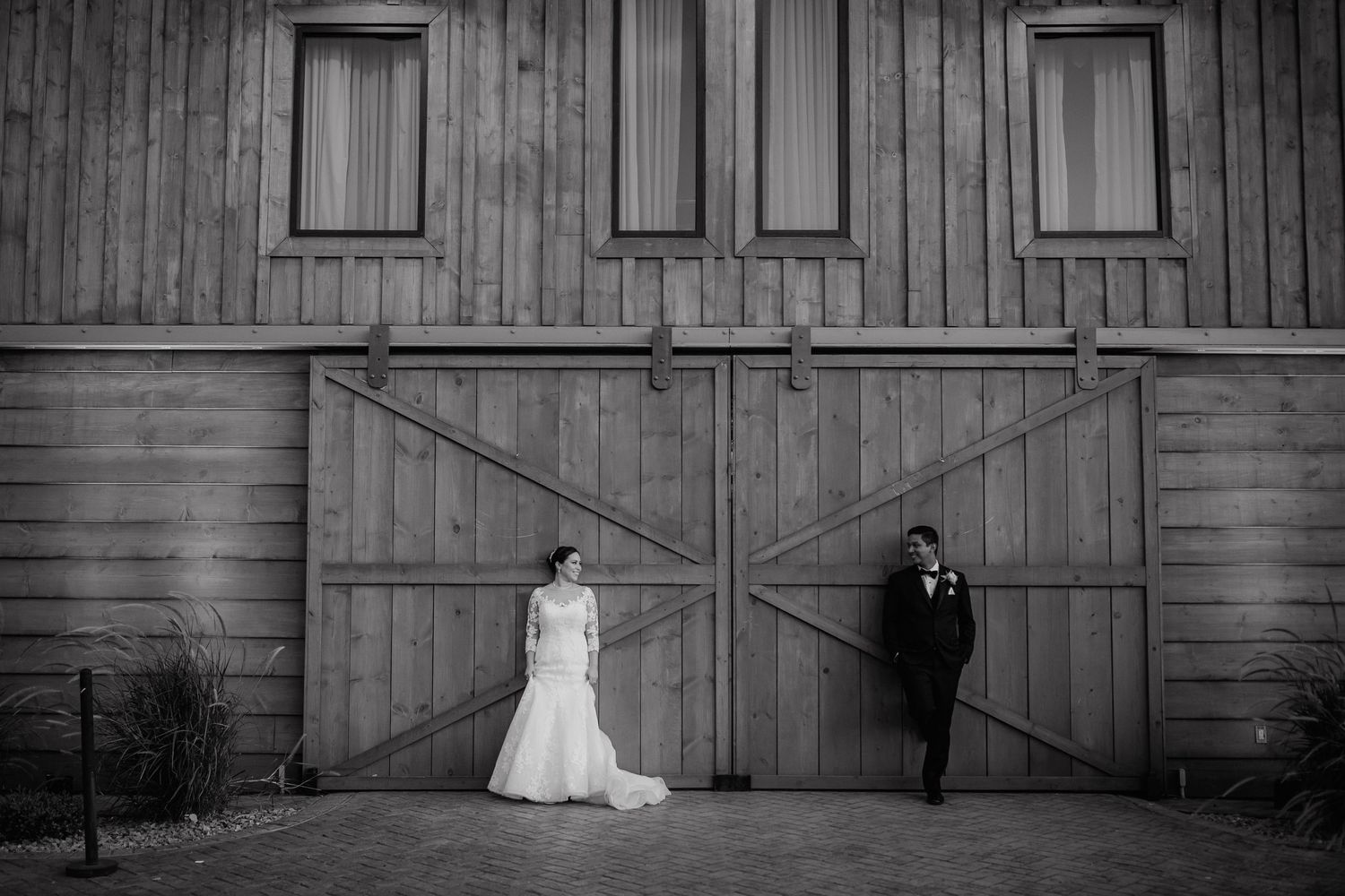 Black and white photo of bride and groom standing apart at rustic barn doors.
