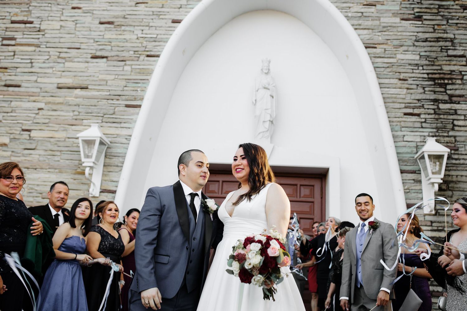 Bride and groom exit church ceremony as guests celebrate with ribbon wands outside.