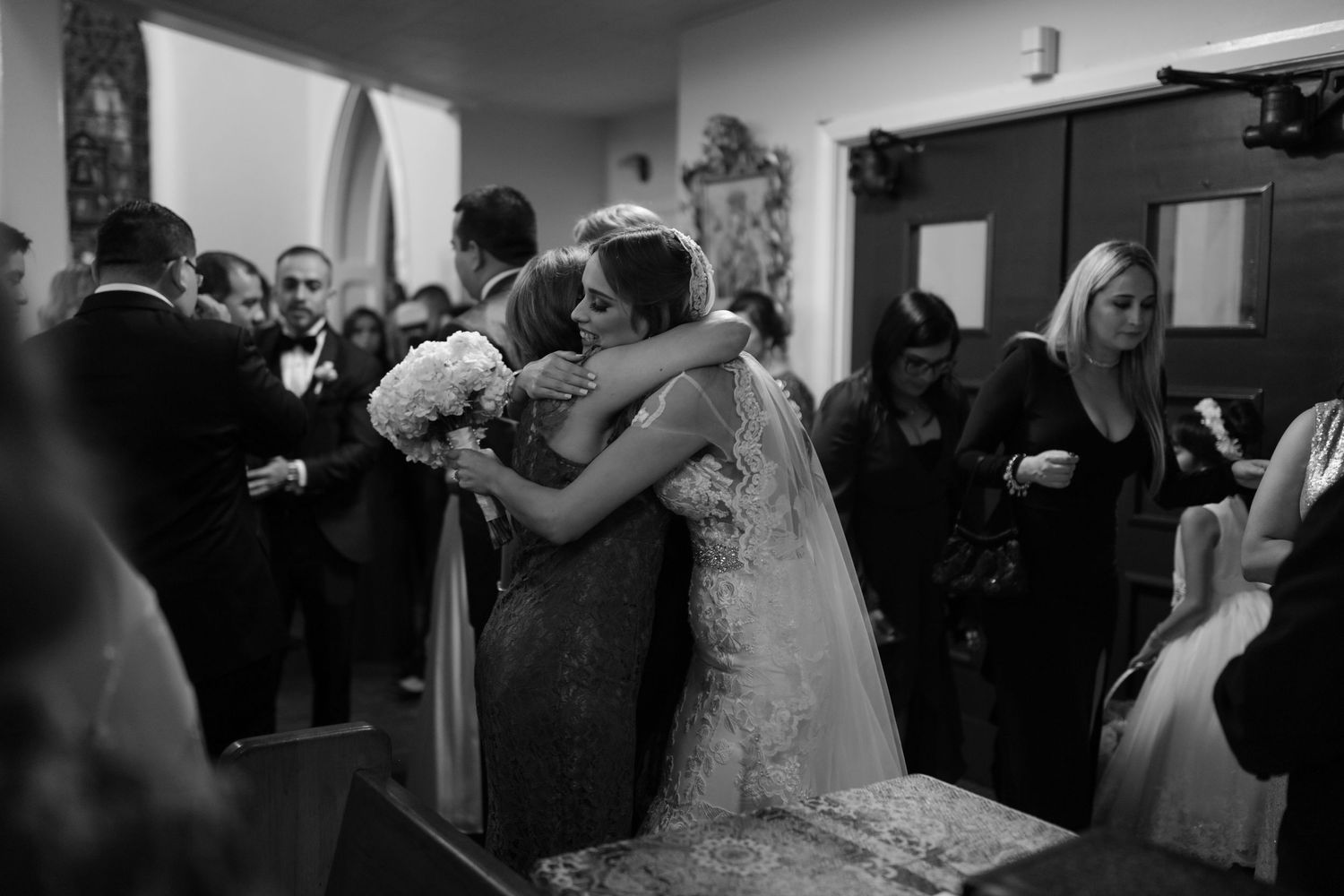 Bride and guest share an emotional hug inside a church during a black and white wedding ceremony.