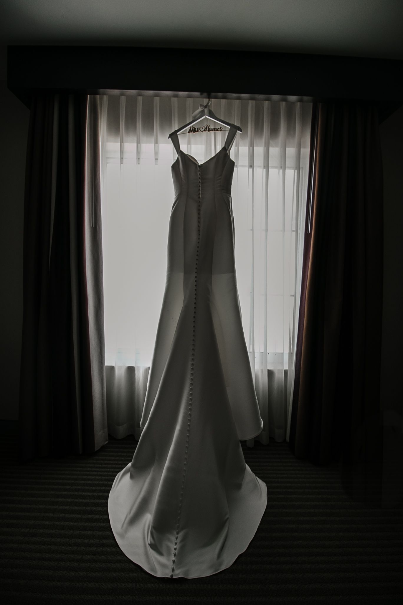 Elegant white mermaid wedding gown hanging on a hanger in front of a window with soft light.