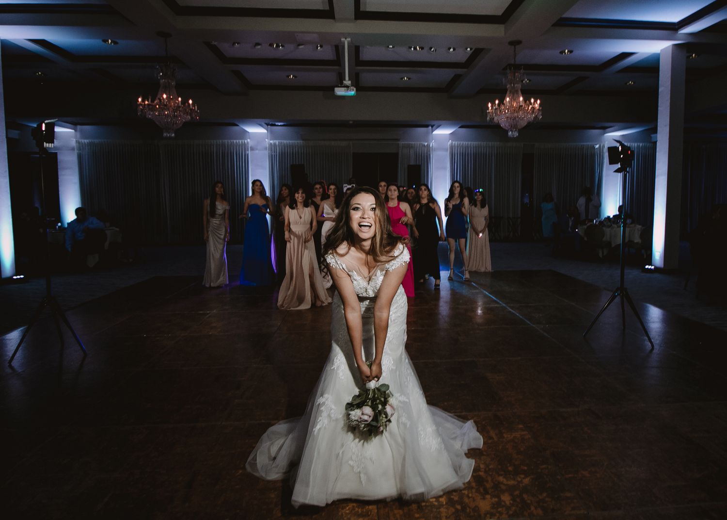 Bride in white tulle gown tosses bouquet to guests gathered in elegant ballroom reception.