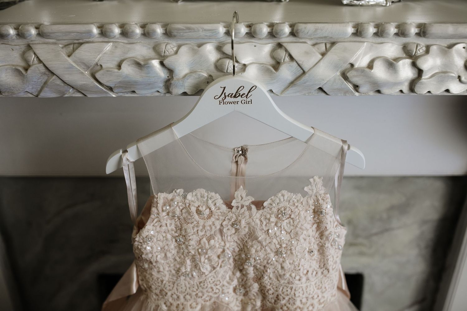 Elegant lace bridal gown hanging on personalized white hanger above ornate fireplace mantel.