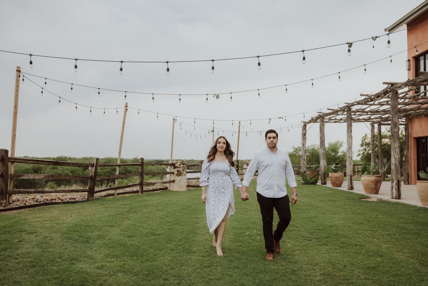 Couple holding hands walking on green lawn with string lights and pergola at outdoor venue.