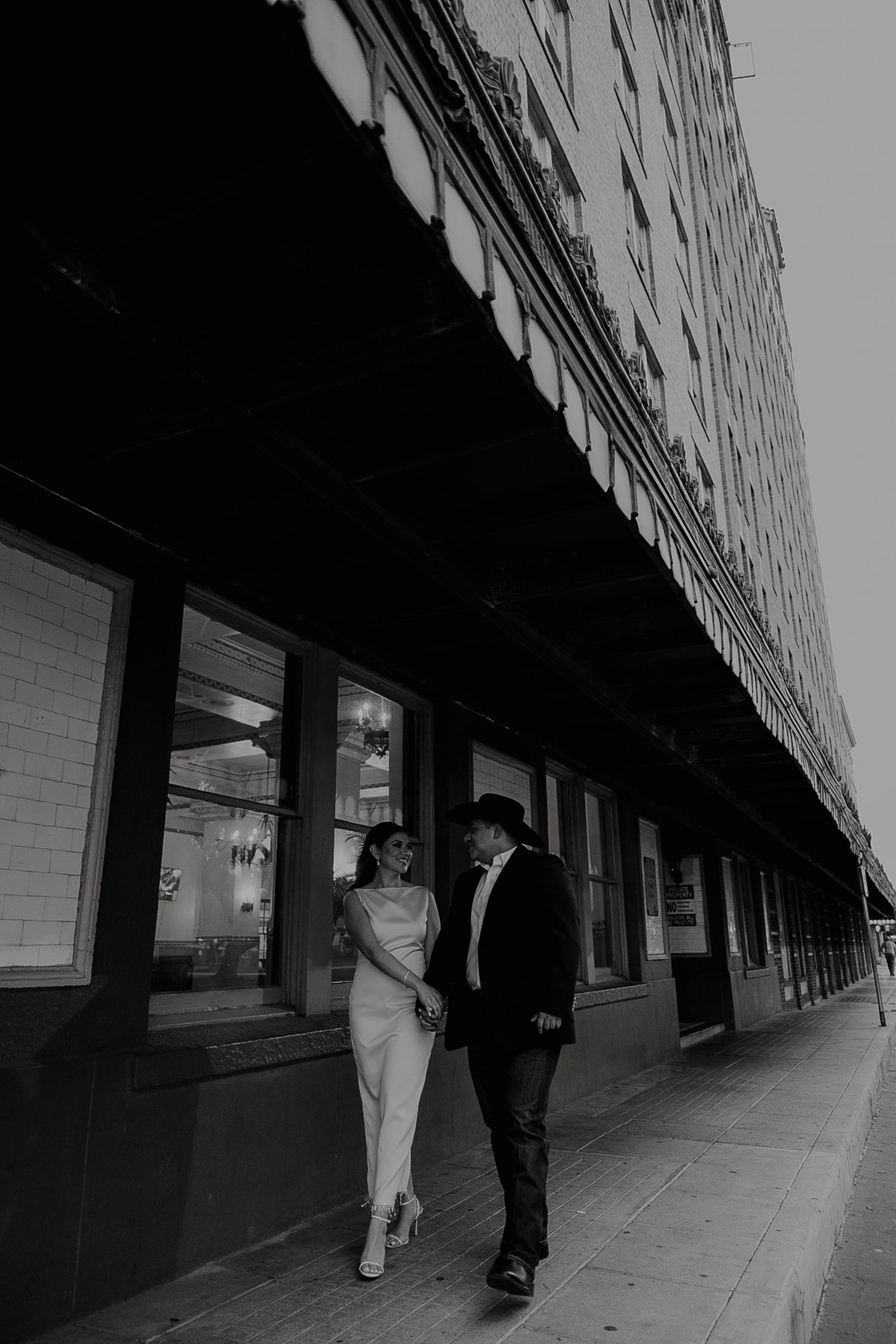 Black and white photo of a couple walking hand in hand along an urban city sidewalk.