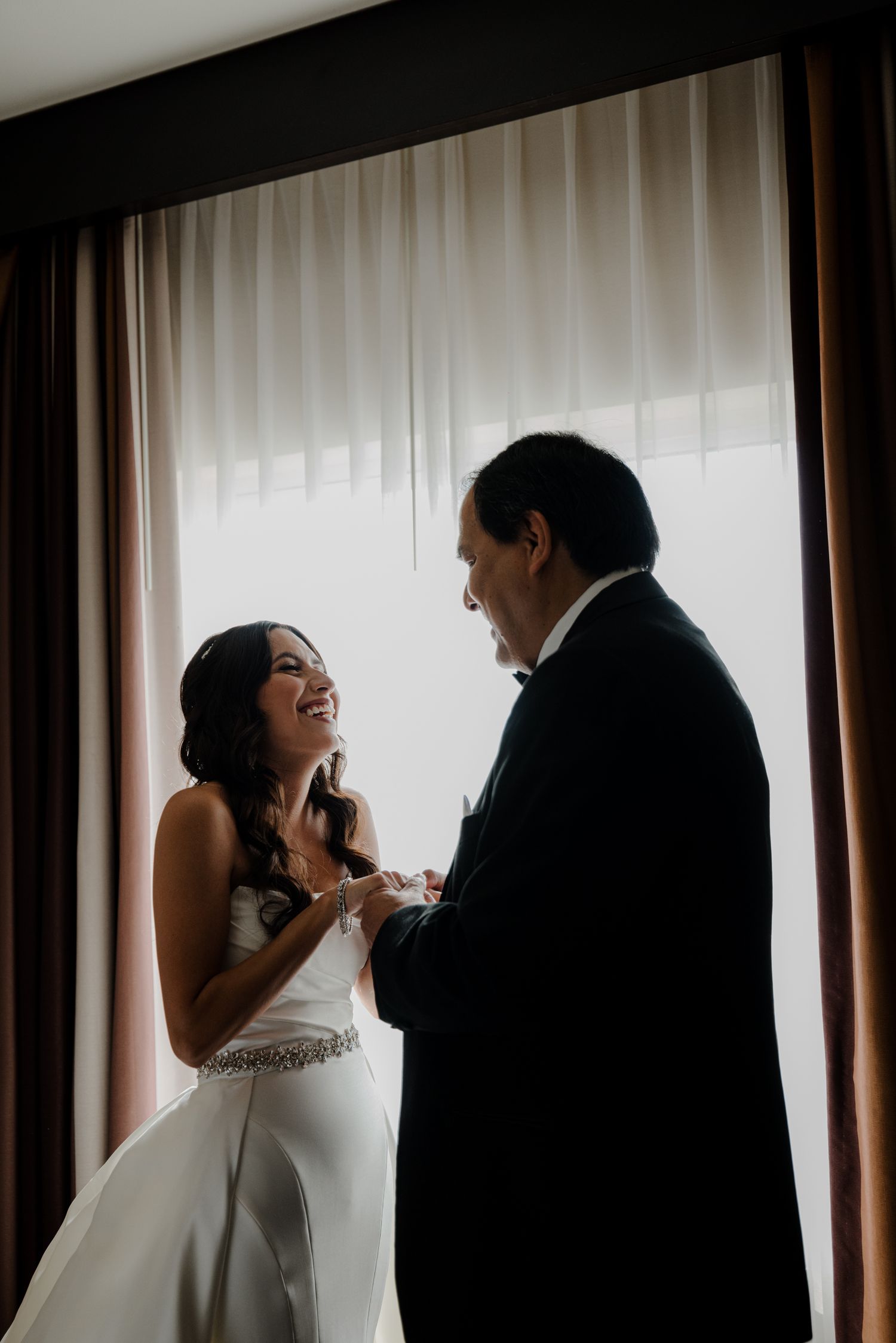 Bride in white gown laughing with groom in tuxedo holding hands by a sunlit window.