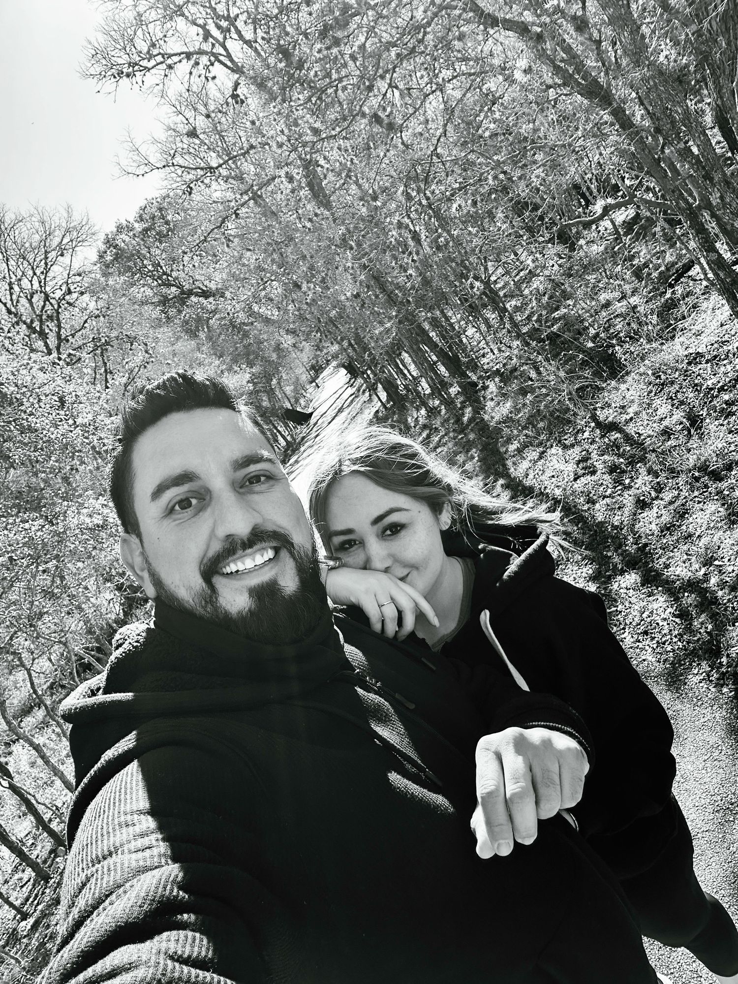 A black and white selfie of a couple smiling together on a path with snow-covered trees in the background.