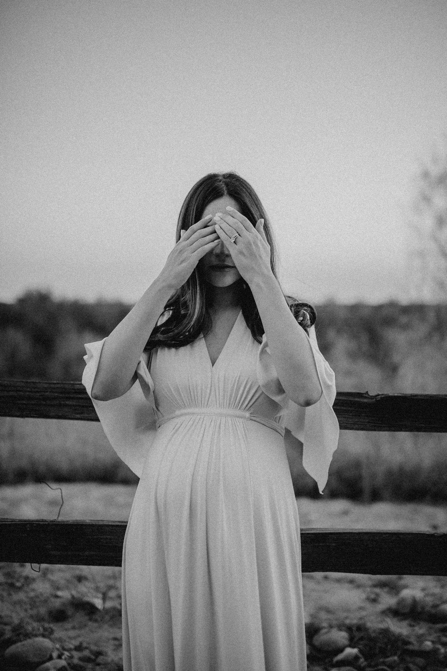 A pregnant woman in white dress covers her face with hands while standing outdoors.