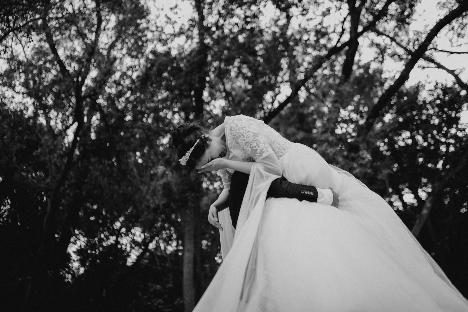 Black and white photo of groom dipping bride in wedding dress outdoors among trees.