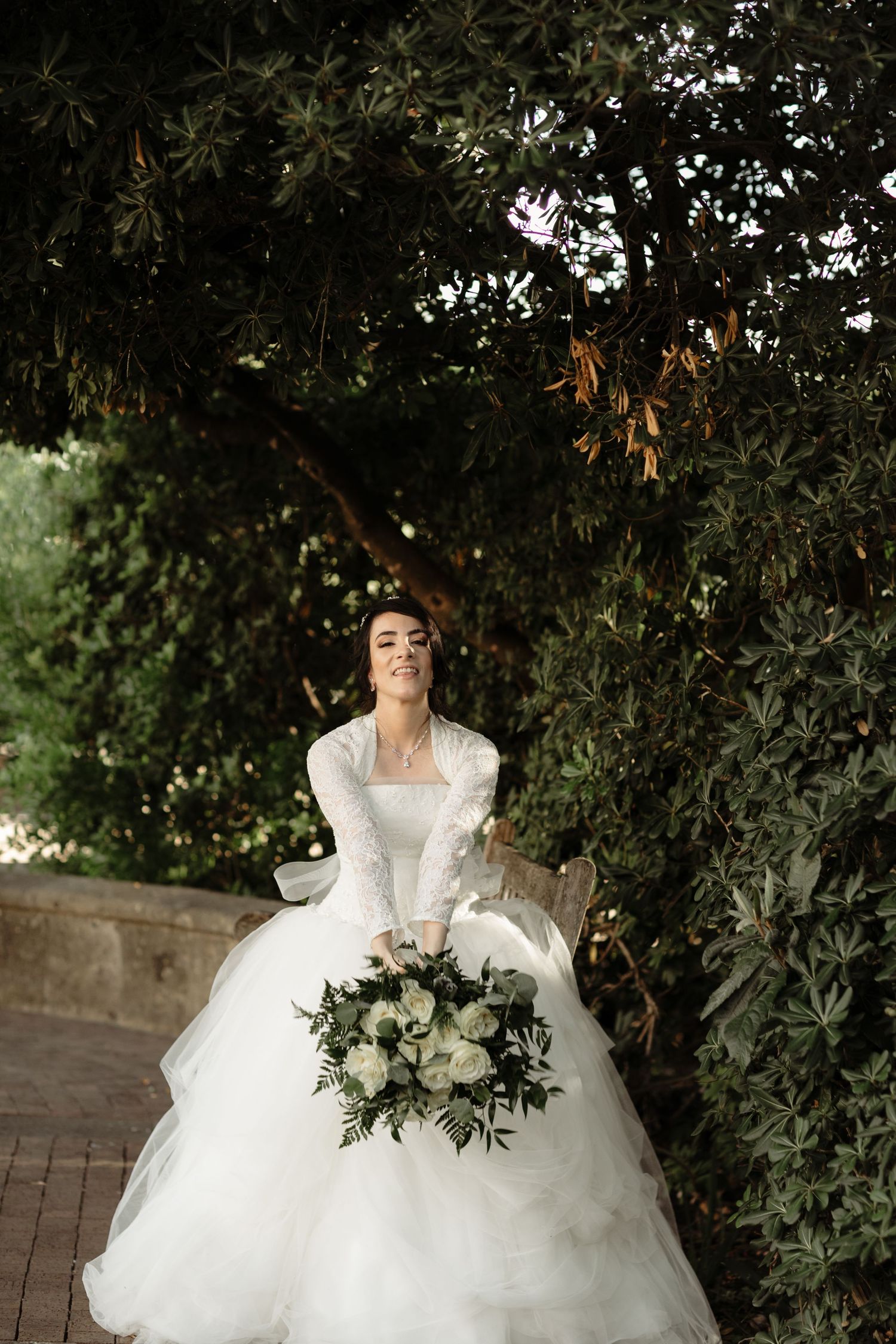 Bride in a white ballgown holding a white floral bouquet, smiling outdoors surrounded by lush green trees.