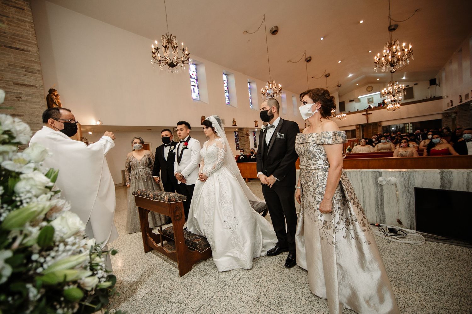 Bride and groom kneel at the altar during a church wedding ceremony surrounded by floral arrangements.