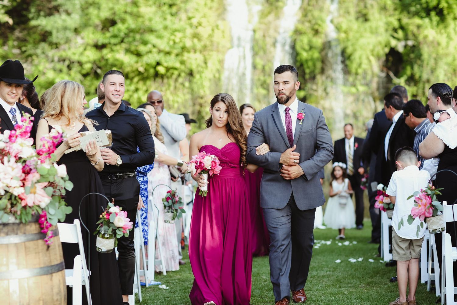 Bridesmaid in fuchsia gown walks down outdoor aisle with groomsman past pink floral arrangements at wedding.