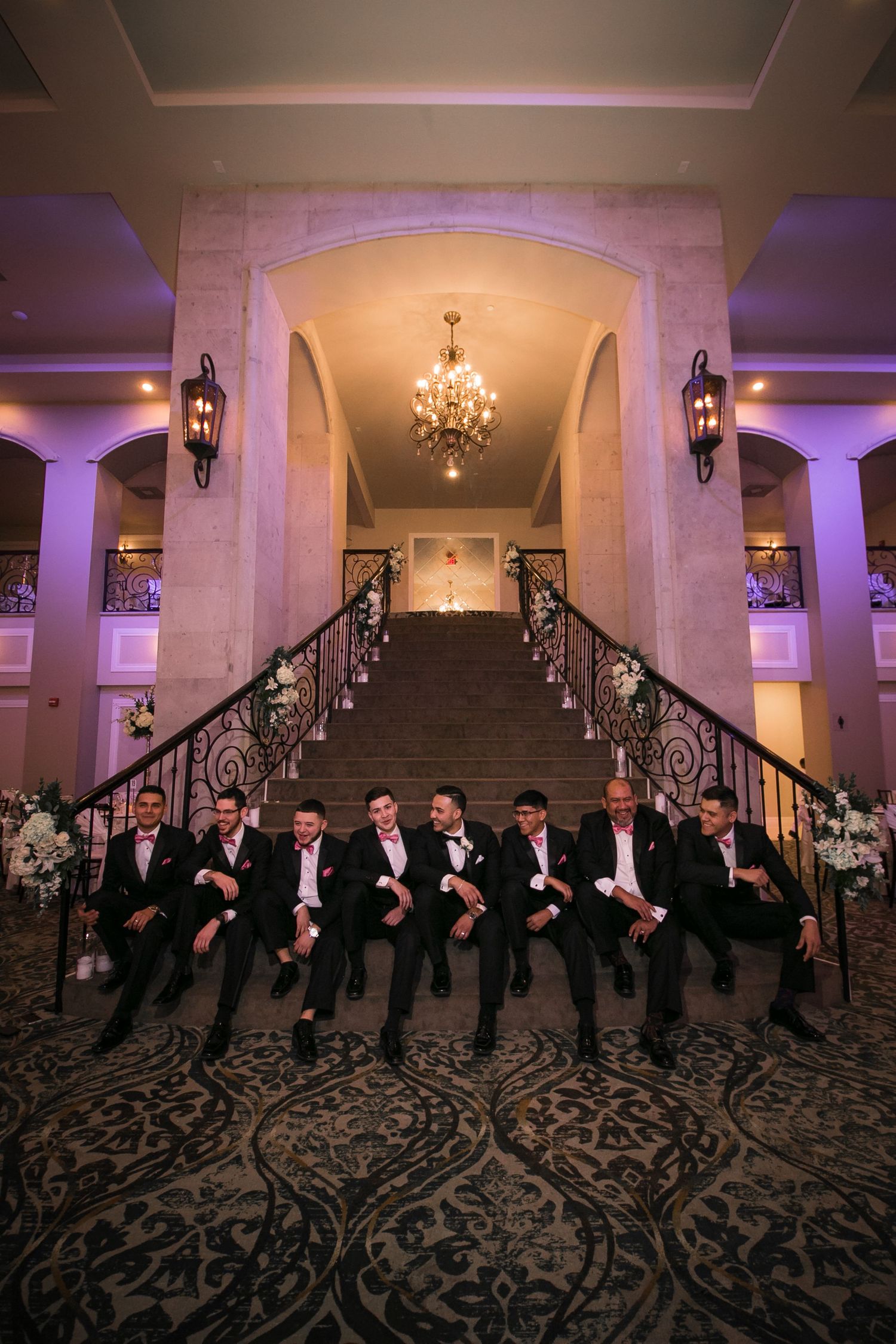 Groomsmen in black tuxedos posing on grand staircase in elegant wedding venue lobby.