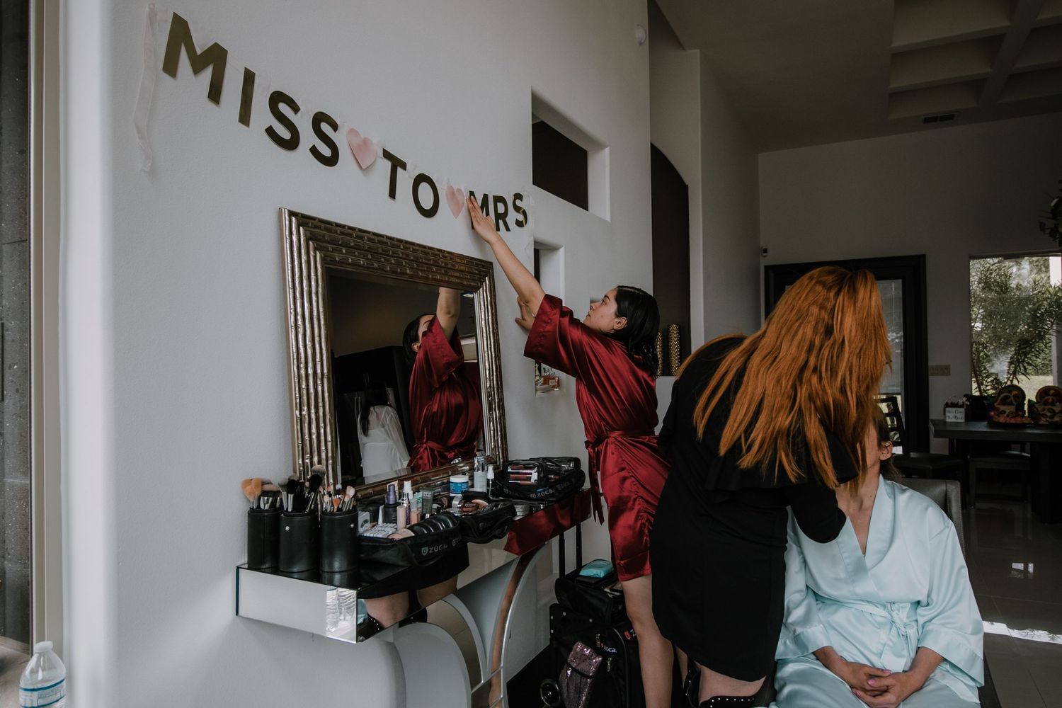 Bridesmaid in red robe hanging a Miss to Mrs banner above a makeup vanity before the wedding.