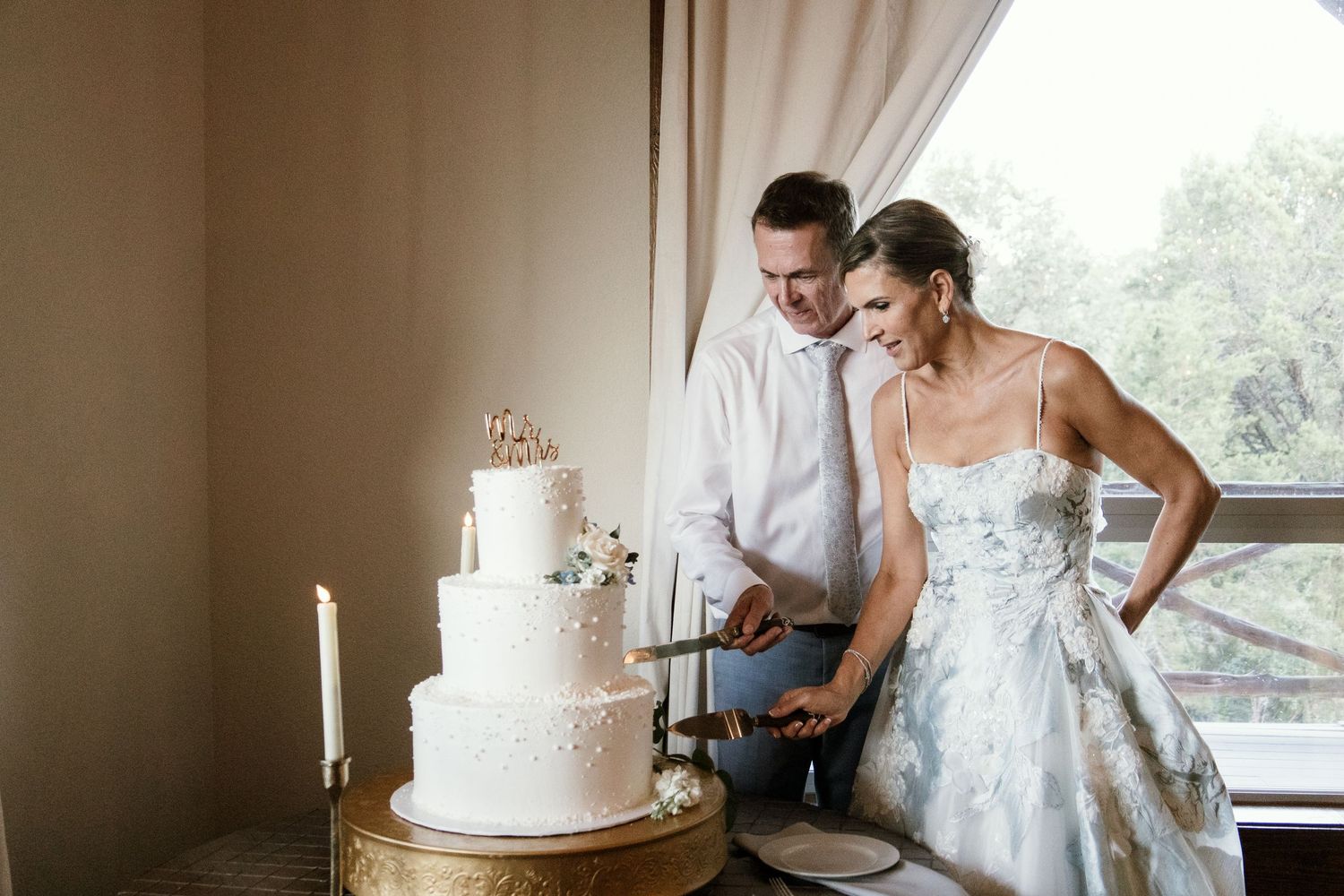 Bride and groom cutting white tiered wedding cake together near window in elegant venue.