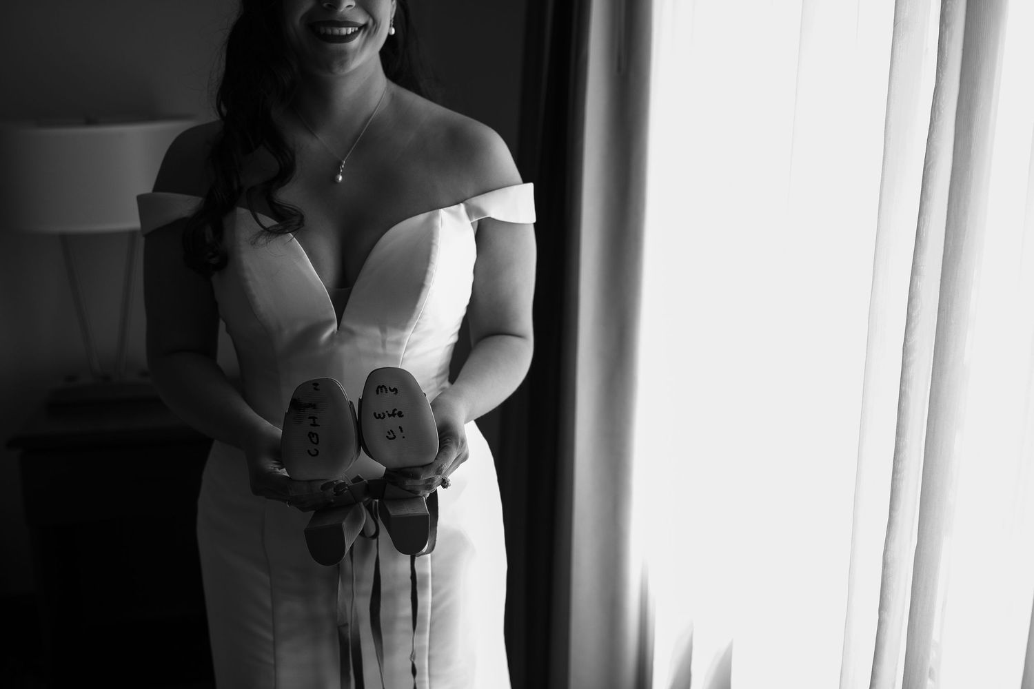 Black and white photo of bride in off-shoulder gown holding shoes with wedding date written on soles.