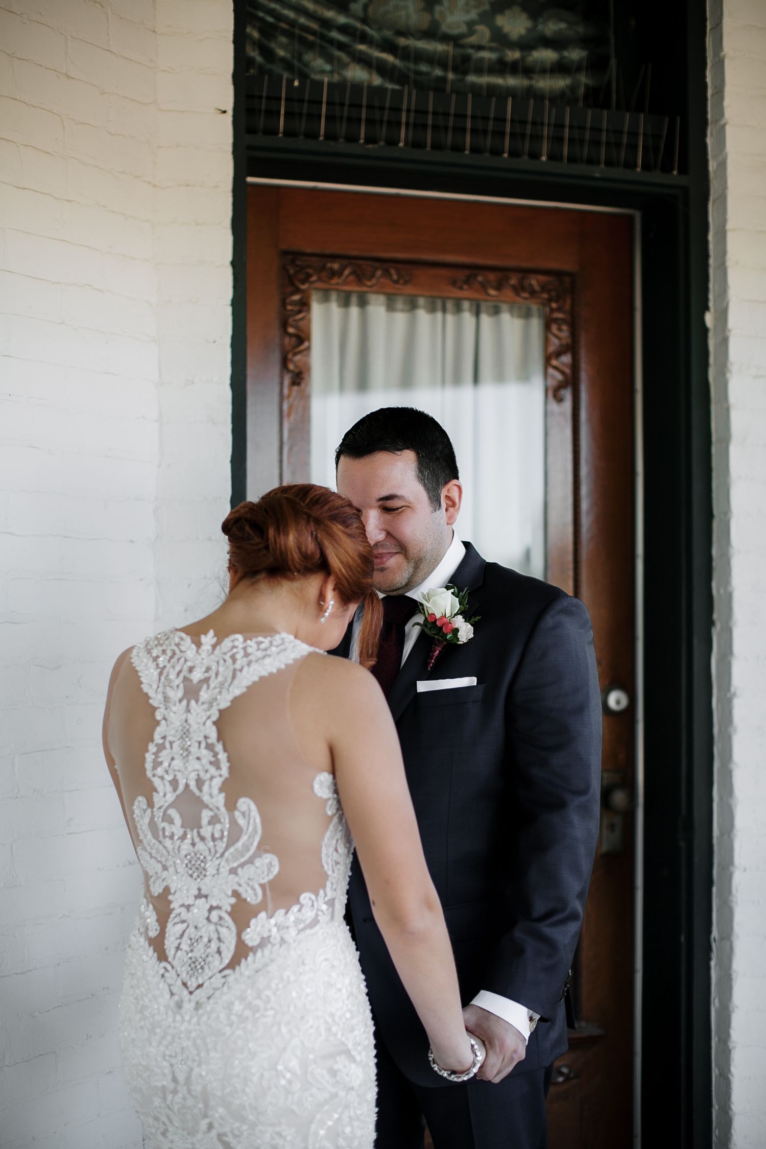 Bride and groom share an intimate moment holding hands in front of an ornate wooden door, showcasing the lace back wedding gown.