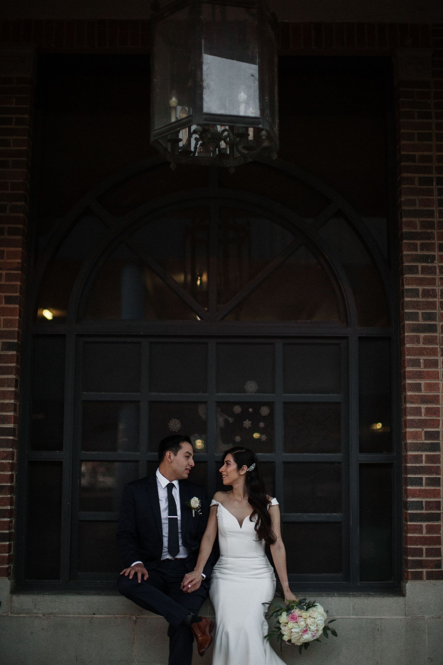 Bride and groom share a romantic moment outside a historic brick building with an ornate lantern overhead.