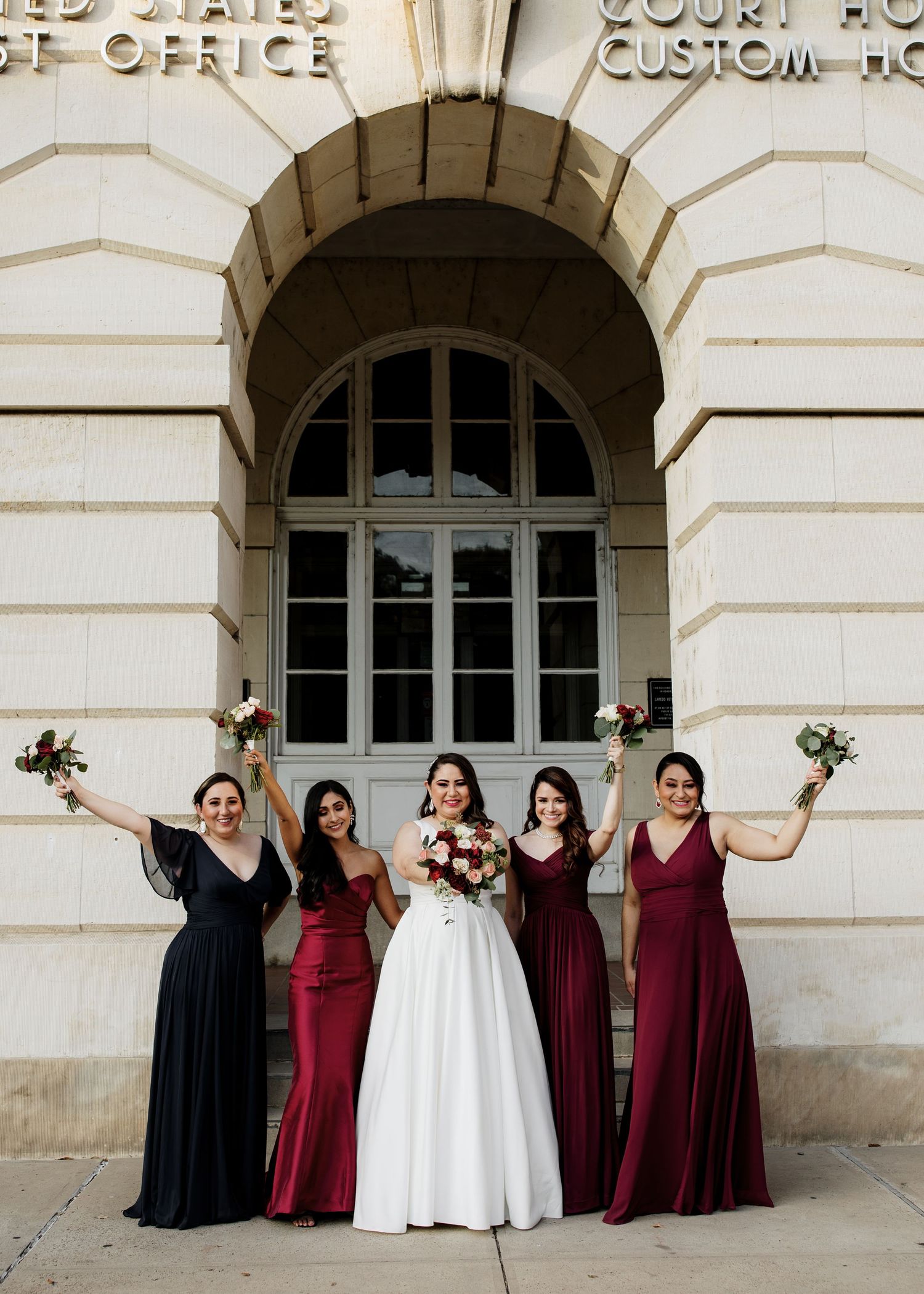 Bride in white gown poses with four bridesmaids in burgundy and black dresses outside historic courthouse.