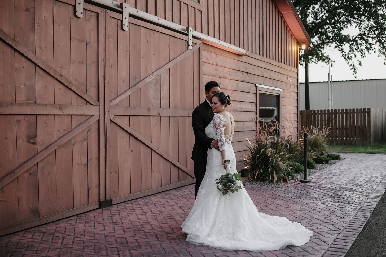 Bride and groom share a romantic embrace in front of a rustic wooden barn door at their wedding.