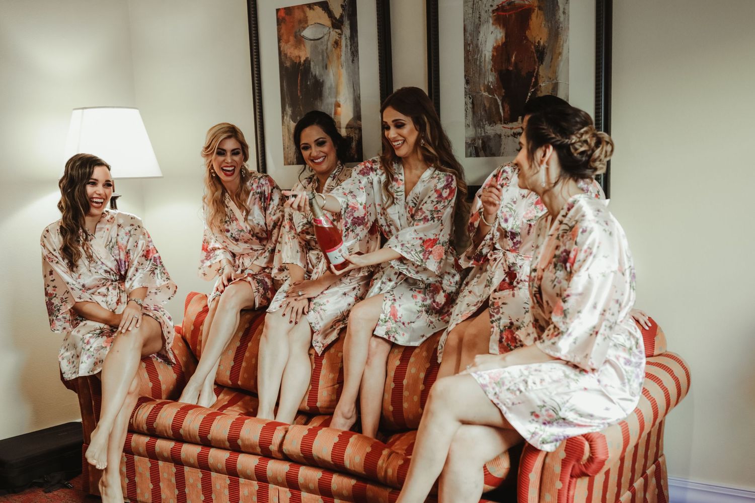 Bridesmaids in floral robes sitting together on sofa during wedding morning preparations.