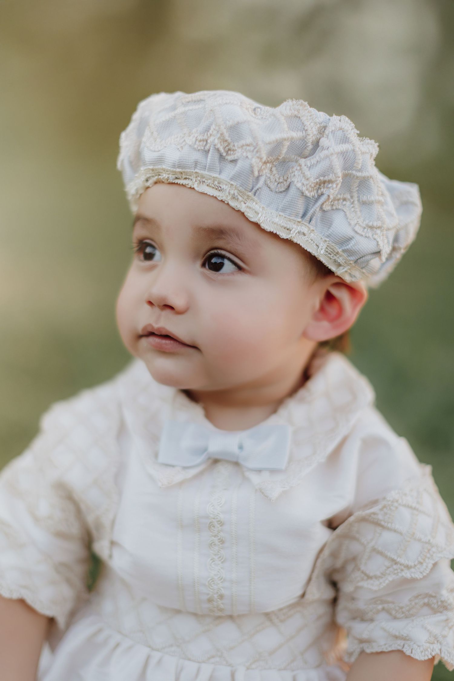 A baby wearing a white vintage-style christening gown and lace bonnet gazes upward in an outdoor portrait setting.