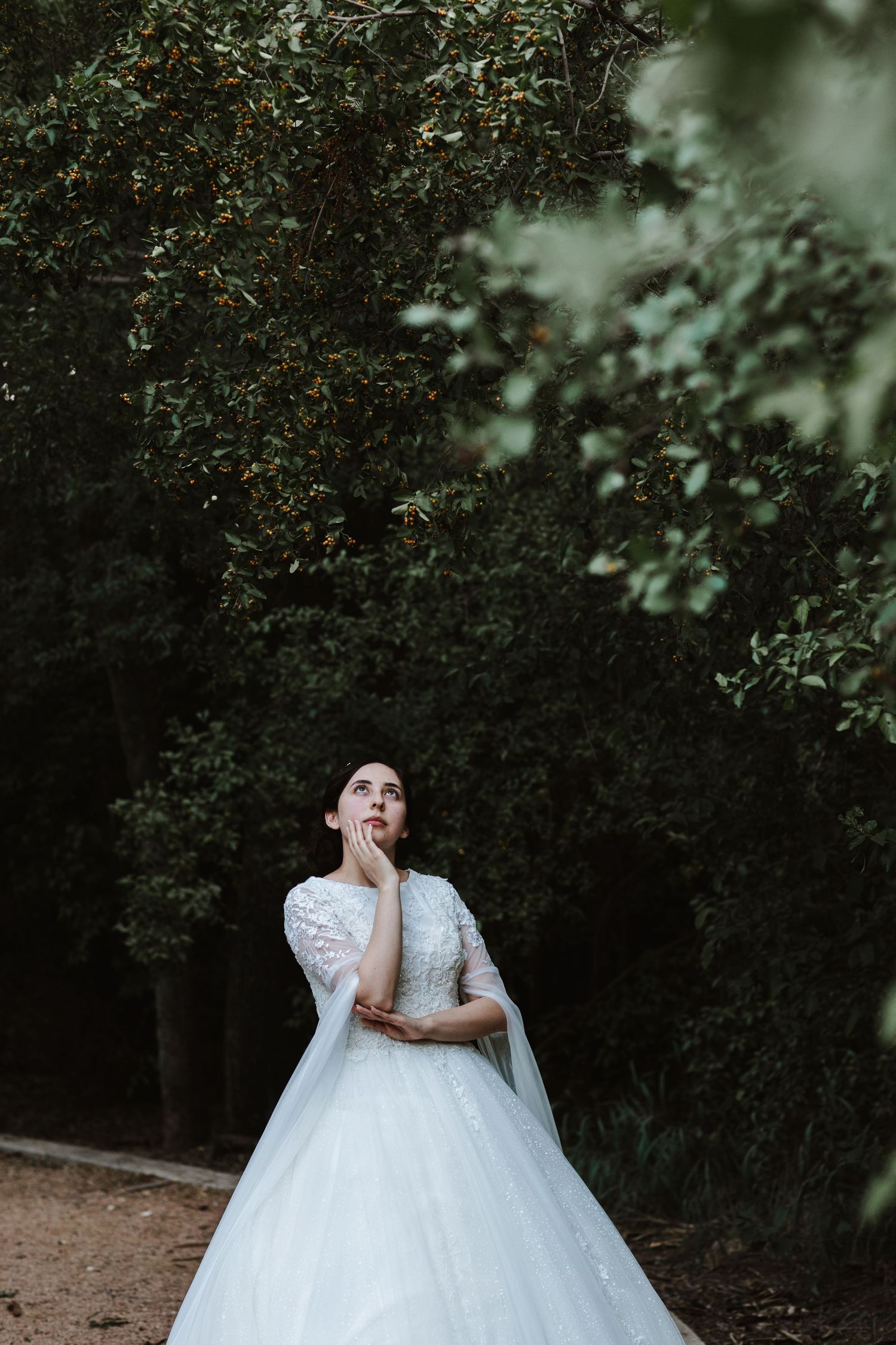 Bride in white ball gown looking up dreamily among lush green trees in an outdoor garden setting.