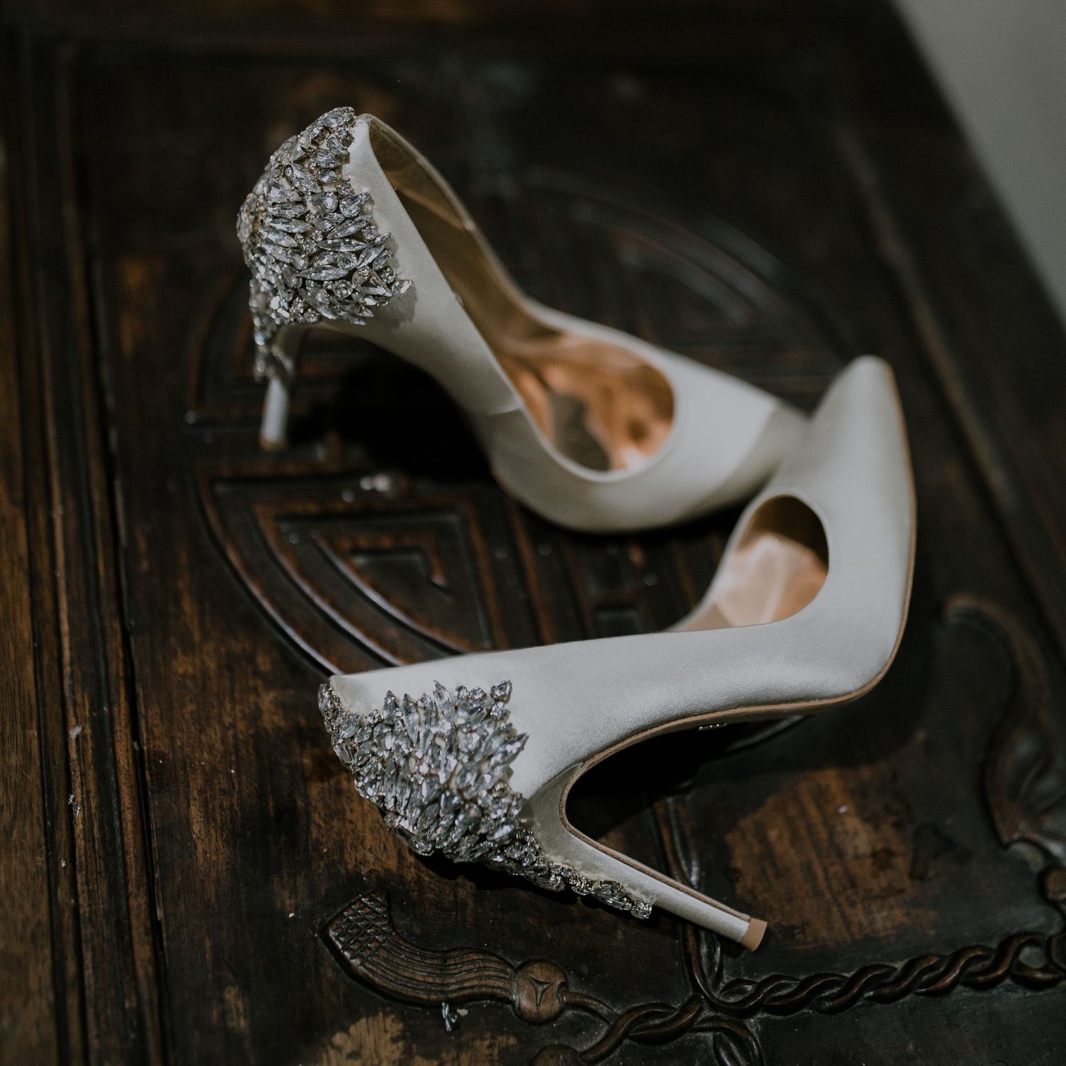 White bridal heels with crystal embellishments displayed on ornate wooden surface.