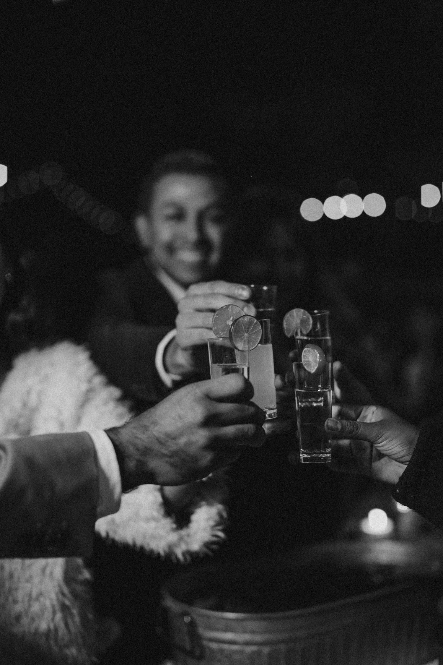 Black and white photo of wedding guests raising shot glasses in celebratory toast at reception.