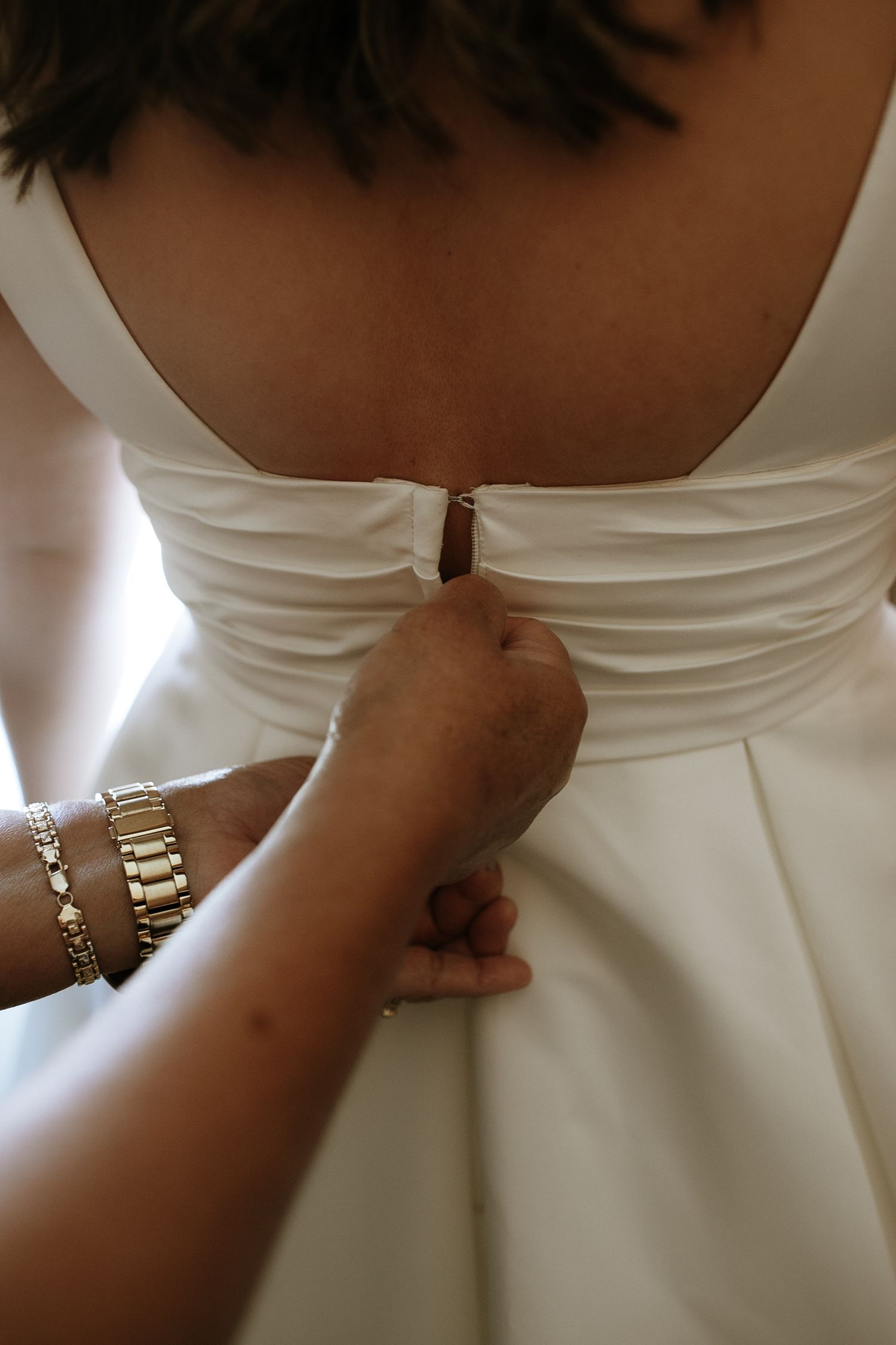 A hand with a gold bracelet fastens the back button of a white ruched wedding gown.