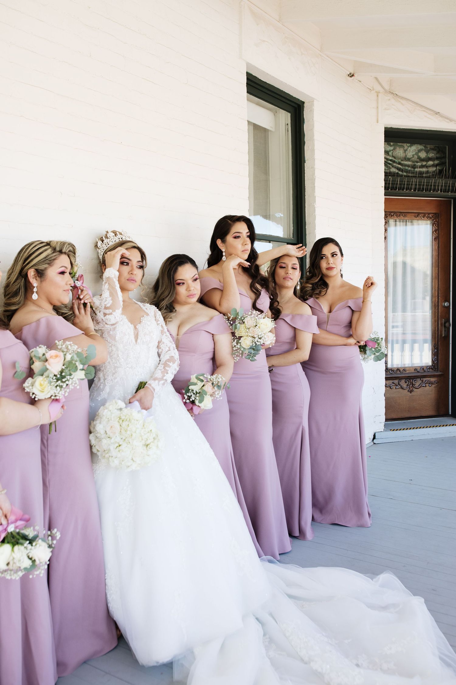 Bride in lace gown with crown poses alongside bridesmaids in lavender dresses holding bouquets.