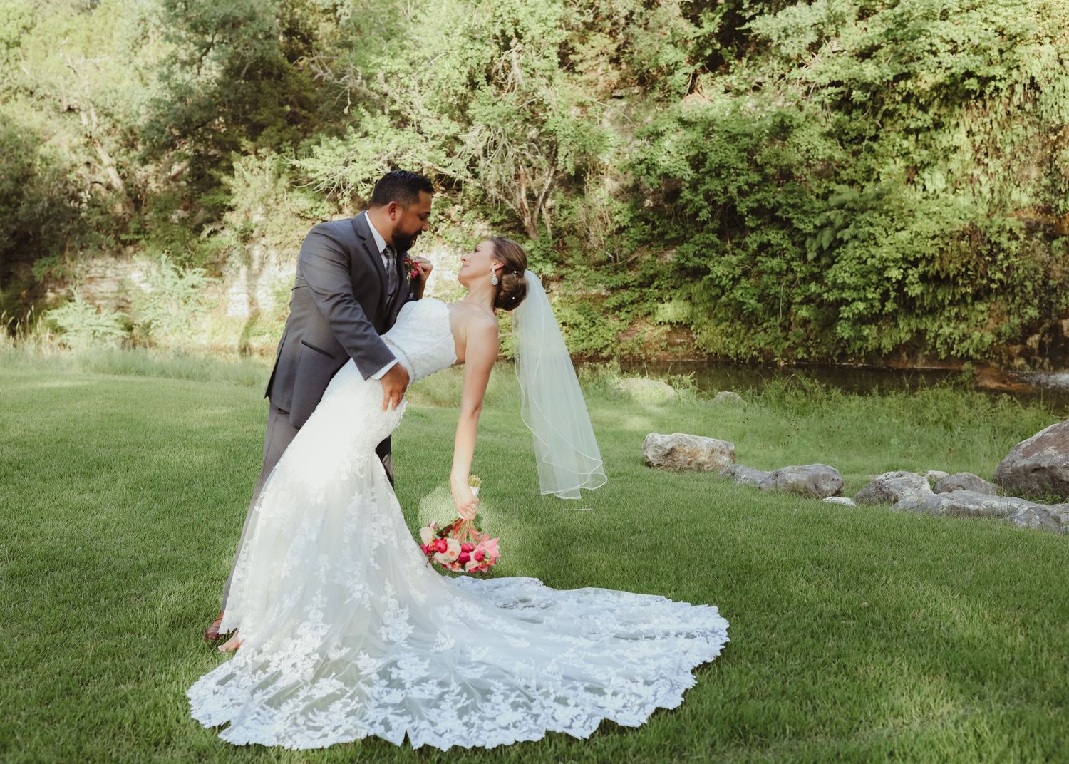 Groom dipping bride in lace wedding gown on green lawn near stream outdoors.