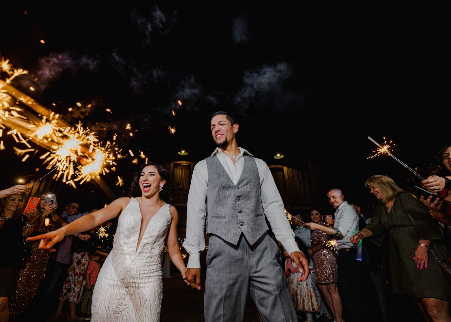 Bride and groom laughing during a sparkler exit surrounded by cheering wedding guests at night.