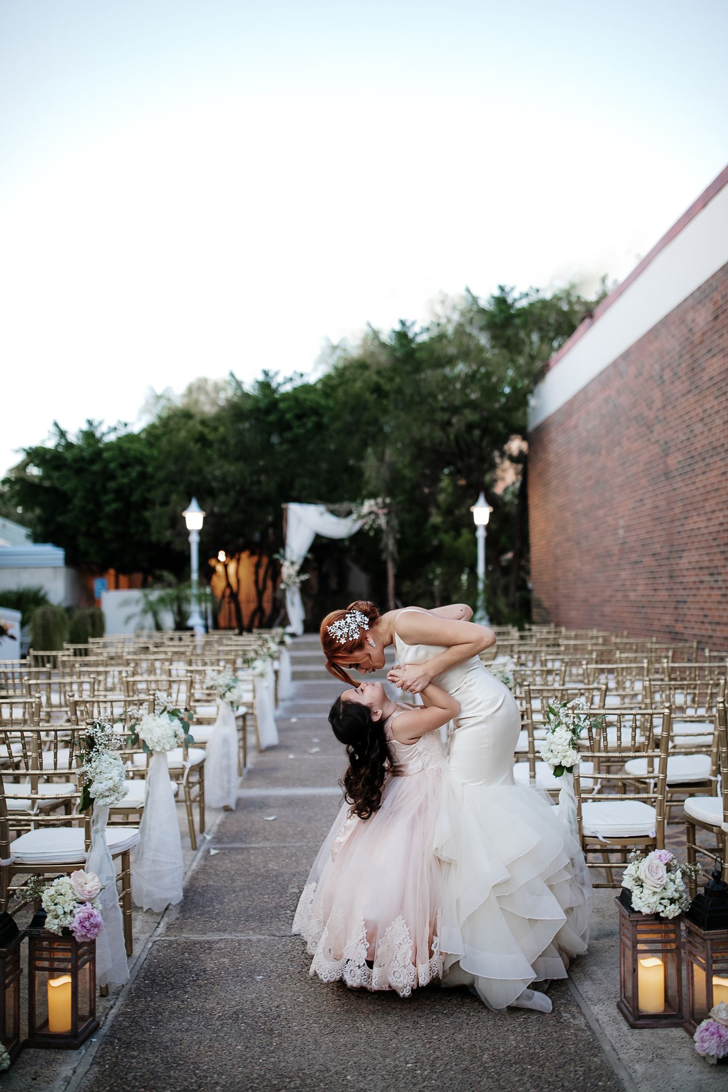 Two brides share a romantic dip kiss at an outdoor wedding ceremony aisle lined with gold chairs and floral arrangements.