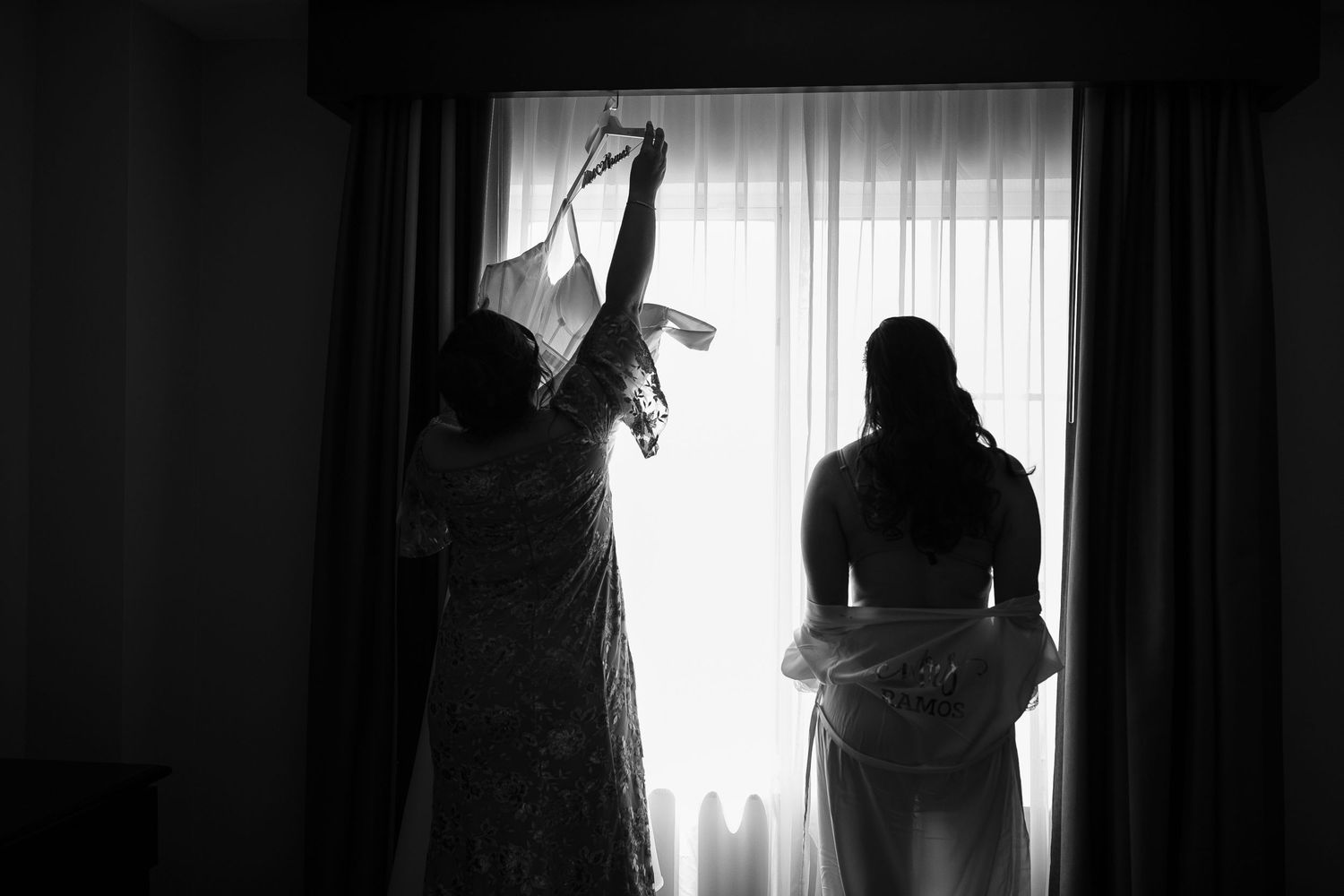 Black and white silhouette of bride and bridesmaid holding wedding dress near bright window before ceremony.