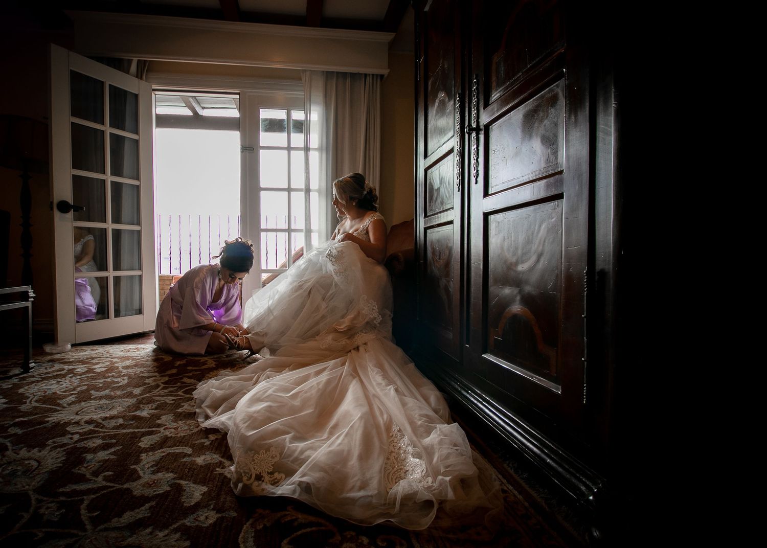 Assistant helps bride with her elaborate white gown in a warmly lit hotel room.