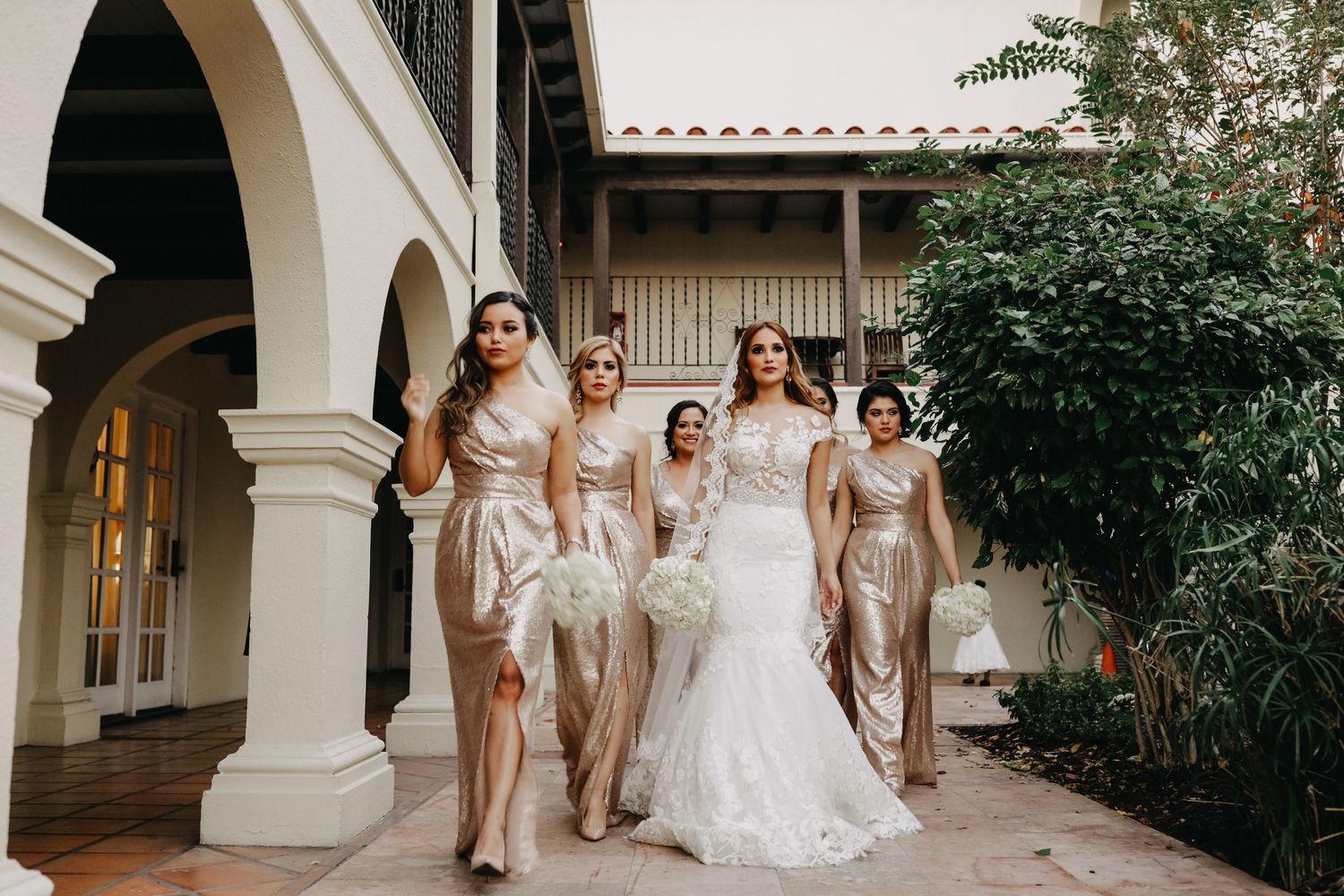 Bride in white gown walks with gold-dressed bridesmaids through elegant courtyard venue.