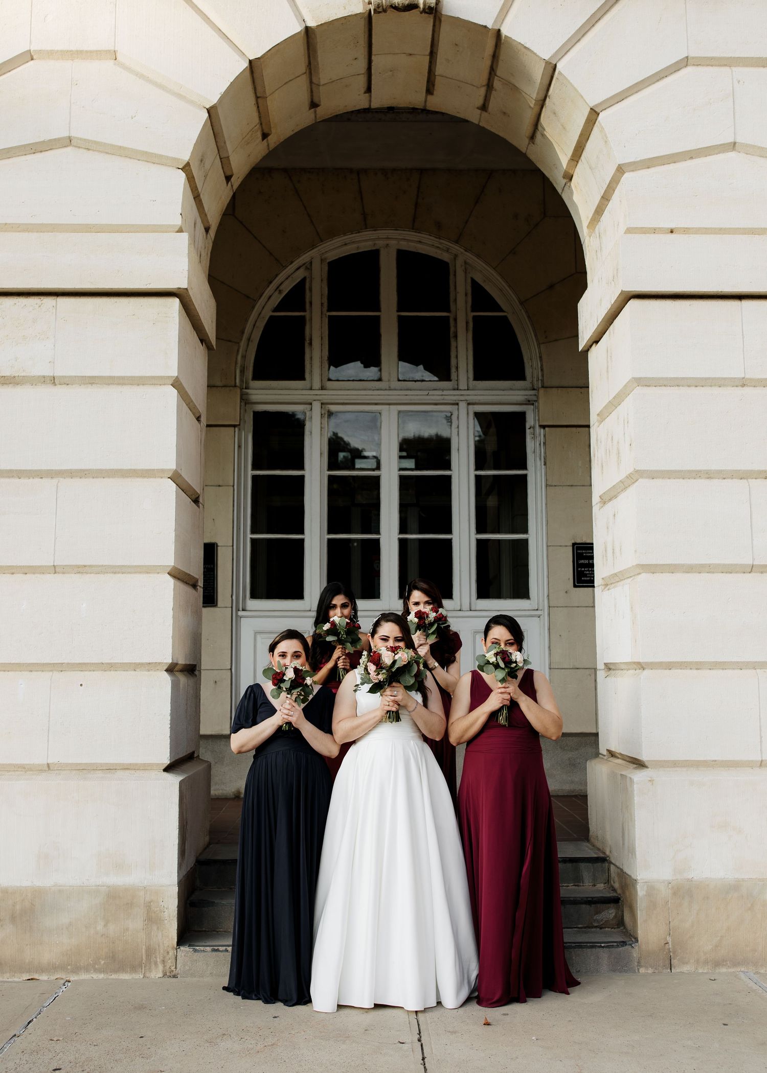 Bride in white gown poses with four bridesmaids in dark dresses holding bouquets before an arched stone building entrance.