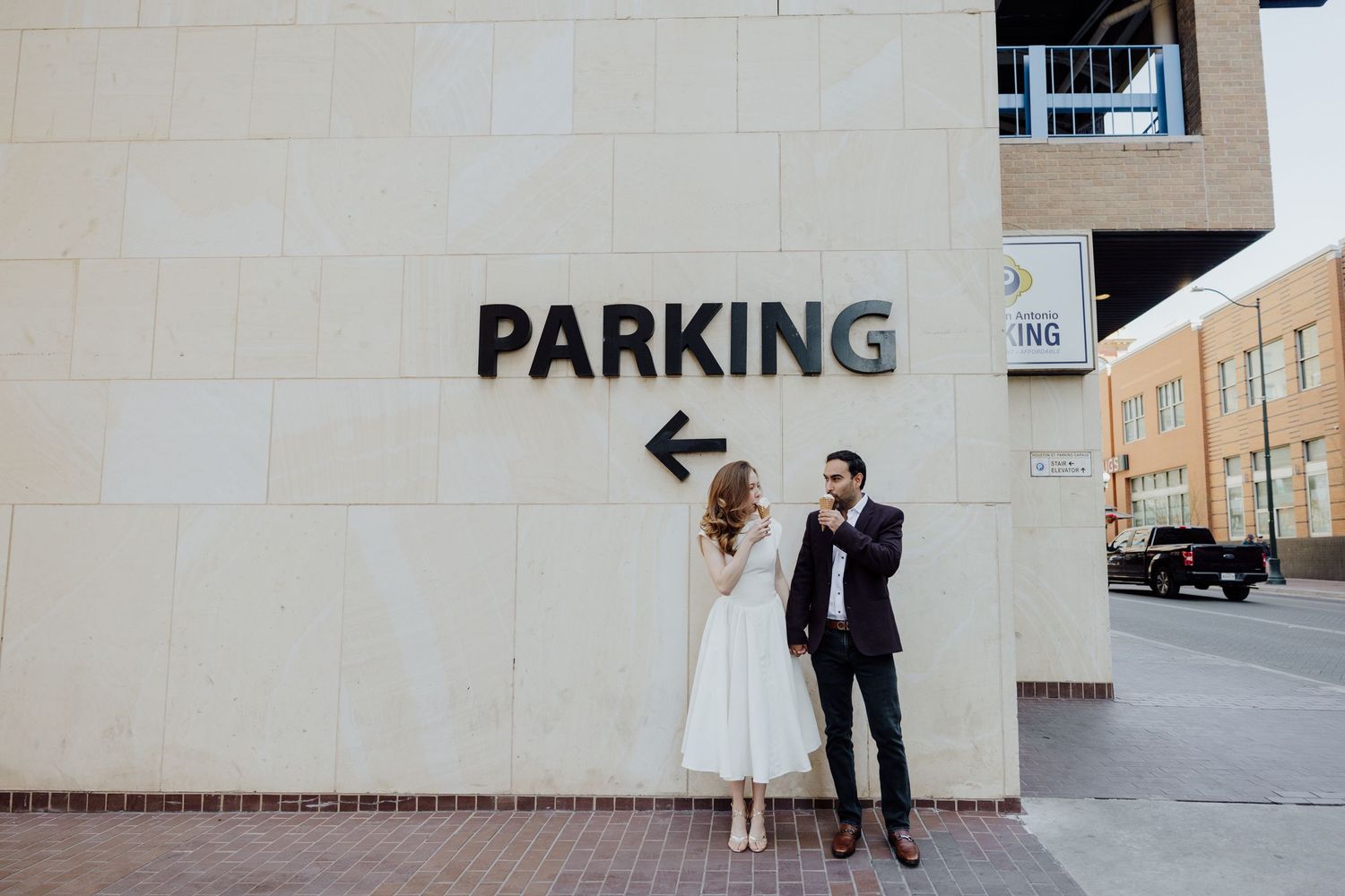 Bride and groom pose by a parking garage sign in an urban setting, bride in white dress, groom in dark suit.