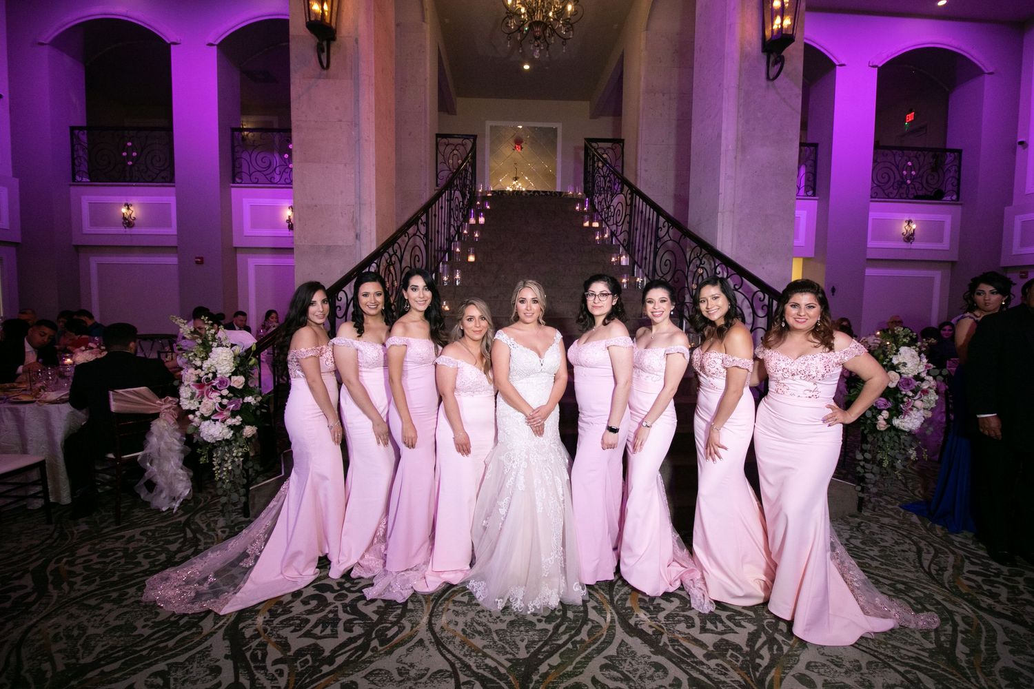 Bride surrounded by ten bridesmaids in pink mermaid gowns posing on grand staircase in ballroom.