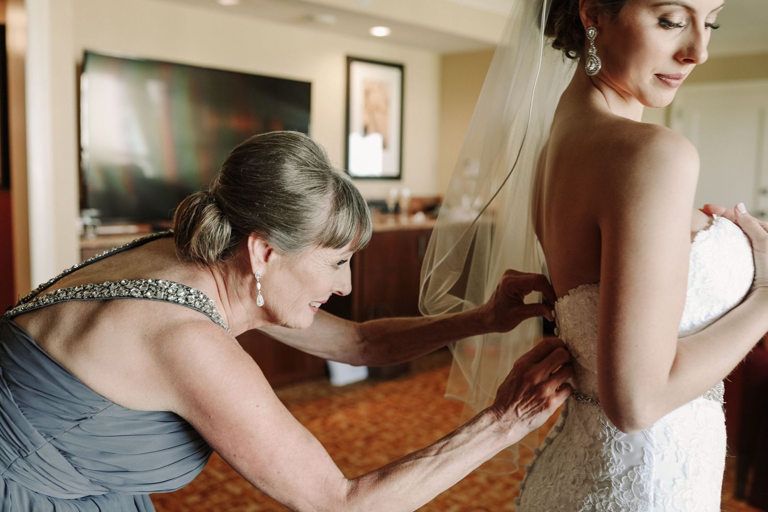 Mother helping bride button up her lace wedding dress during bridal preparations.