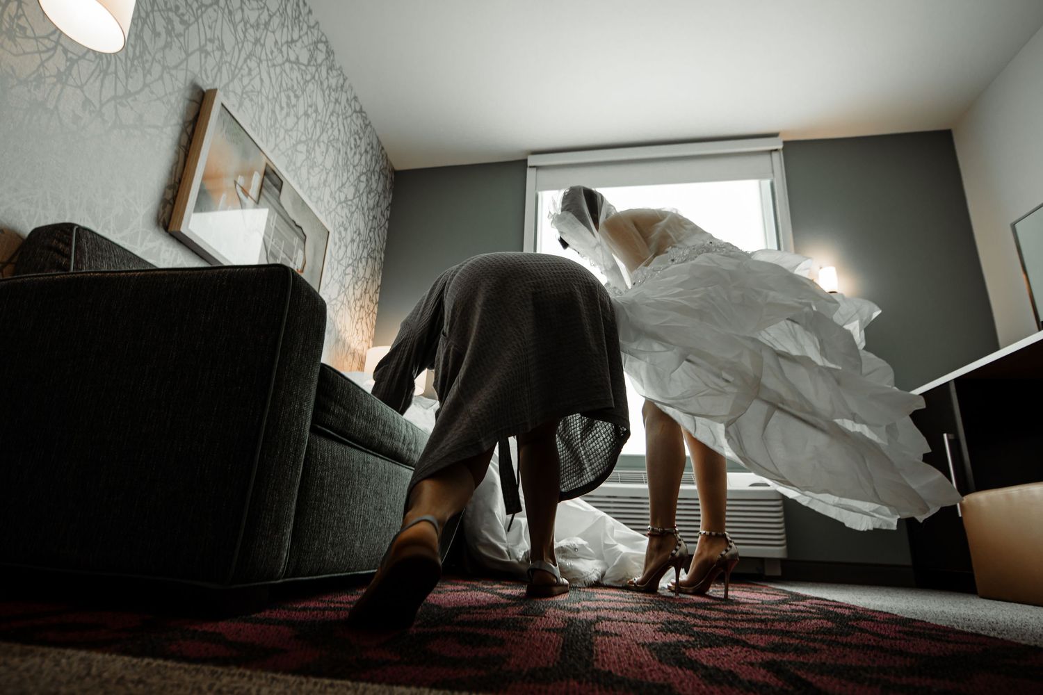 Bride in full white gown being helped with dress in hotel room.