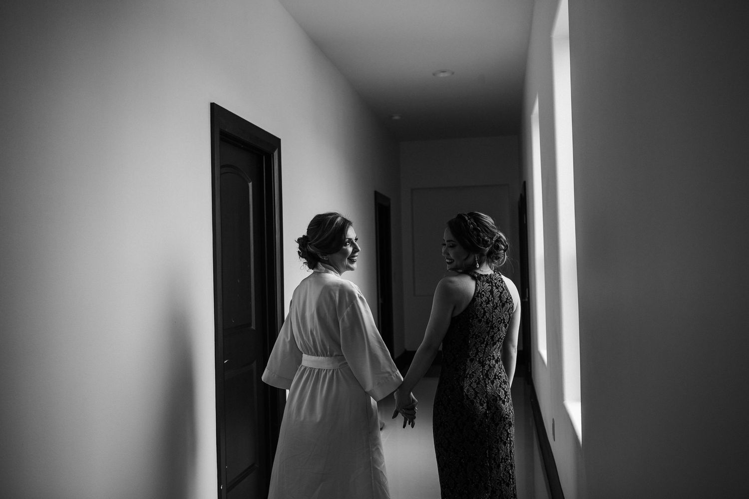 Bride in white robe holds hands with guest in black and white hallway photo before wedding ceremony.