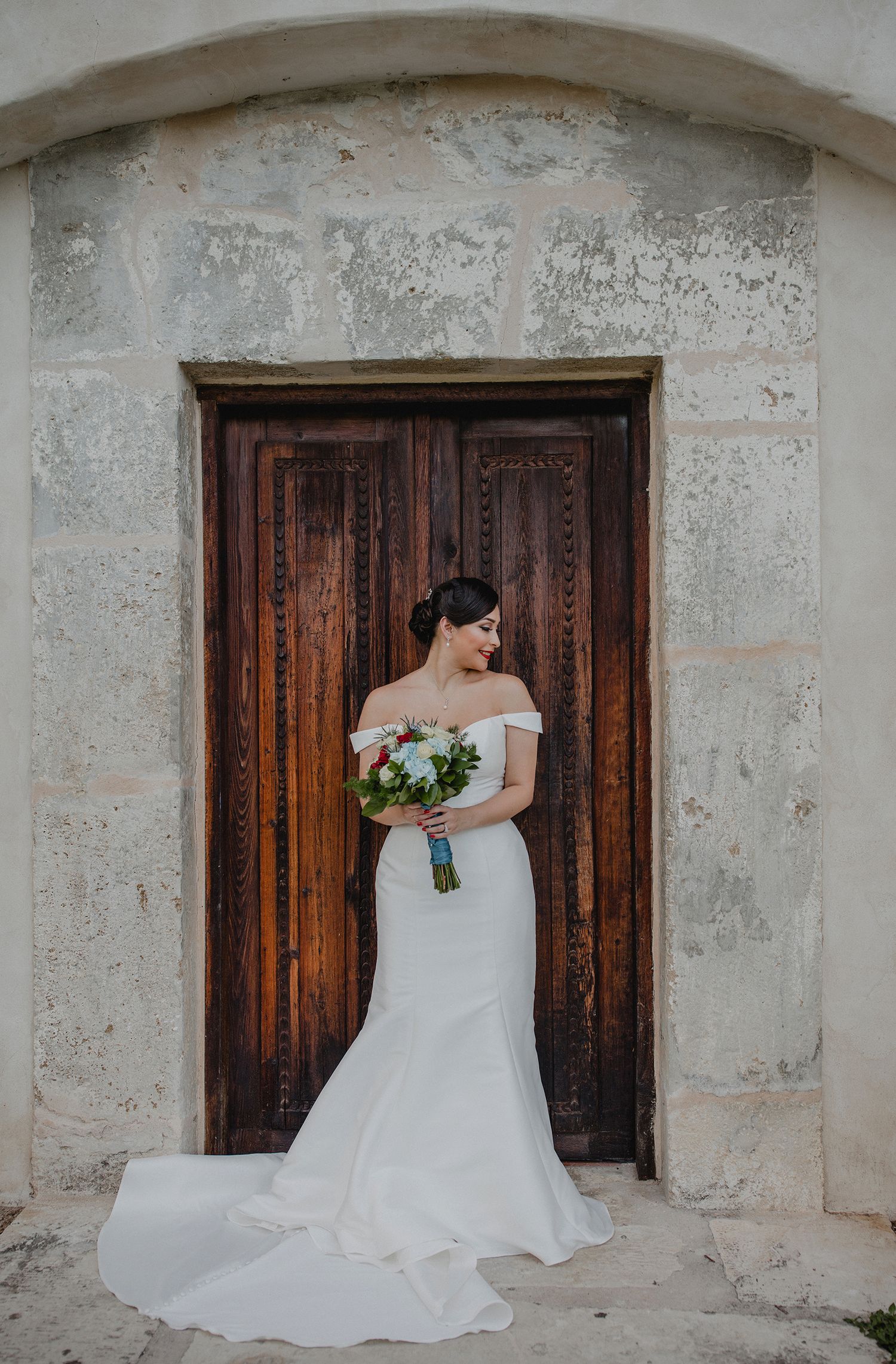 Elegant bride in off-shoulder mermaid gown holding bouquet before ornate wooden church doors.