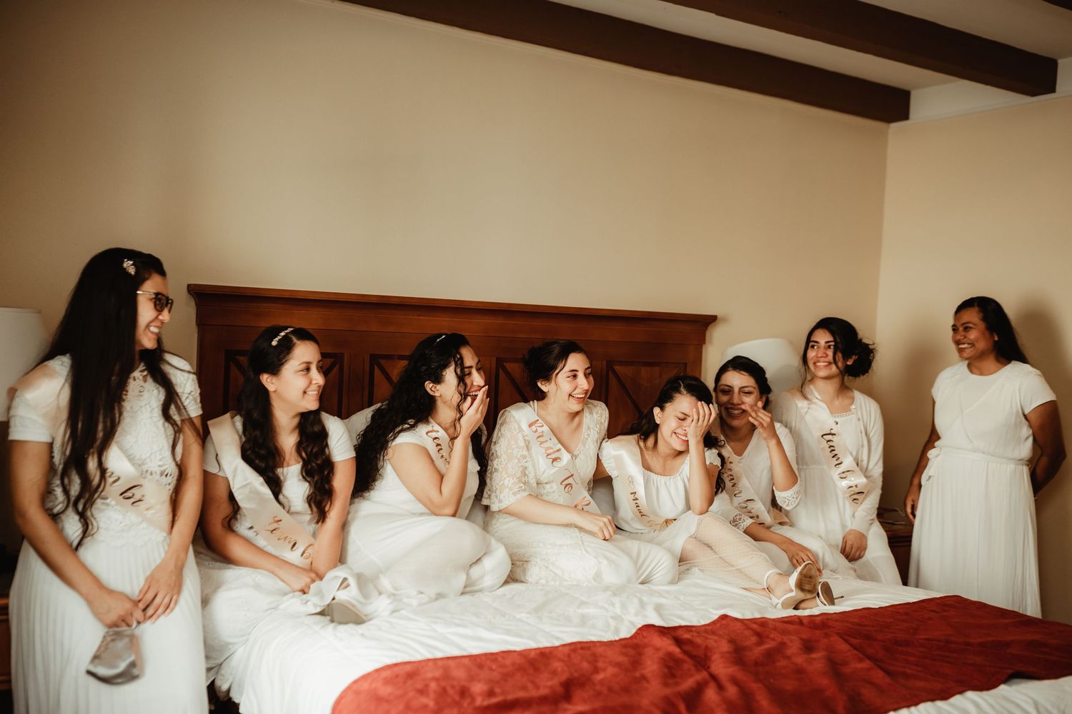 Bridesmaids in white dresses laughing together on a bed during wedding preparations.