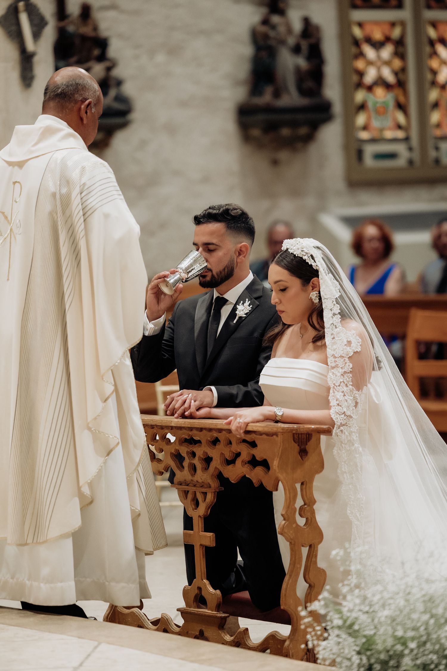 Bride and groom kneeling at altar during Catholic wedding ceremony as priest officiates in white vestments.